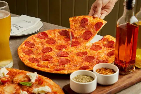 Pepperoni pizza with three small bowls of spices and a glass of beer