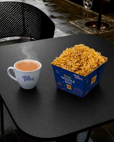 Steaming cup of chai and a box of crispy desi noodles on a black table
