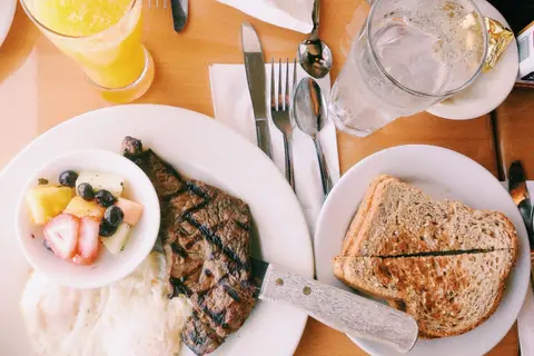 Grilled steak with fried eggs, fruit bowl, toasted sandwich, and drinks