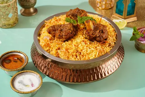 Mutton biryani served in a metal bowl with raita and curry on the side