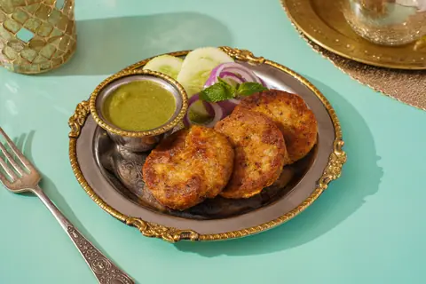 Three pieces of kebab with green chutney, onion, and cucumber slices on a decorative plate